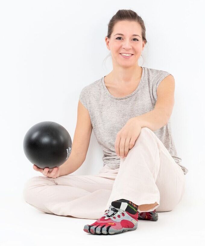 Eine Frau in heller Kleidung und Zehenschuhen sitzt auf dem Boden, lächelt und hält einen schwarzen Ball vor einem weißen Hintergrund.
