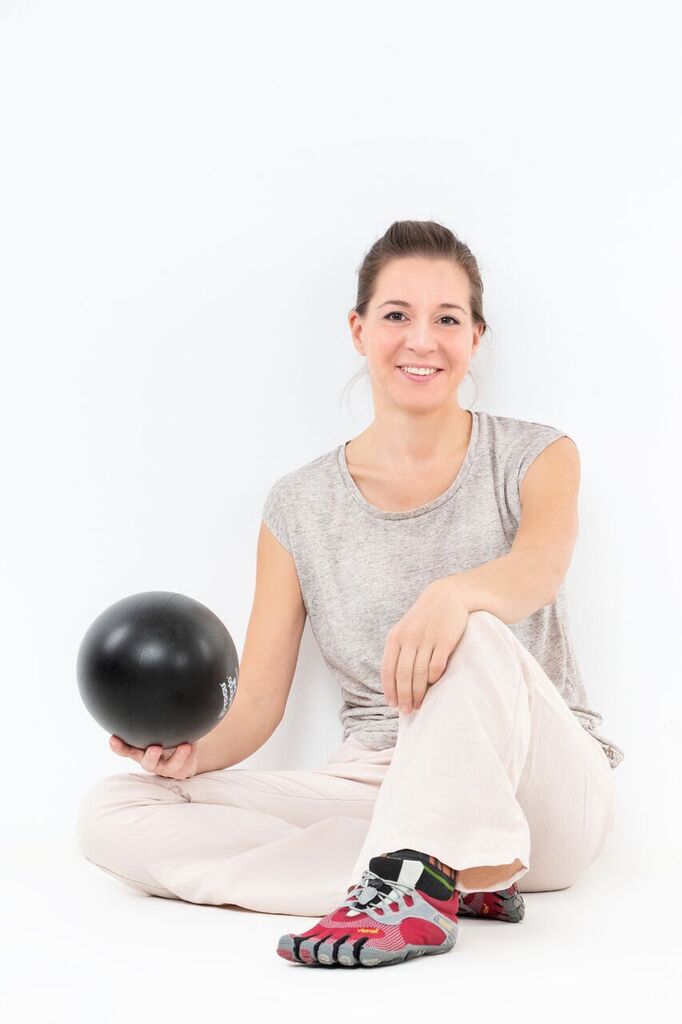Eine Frau in heller Kleidung und Zehenschuhen sitzt auf dem Boden, lächelt und hält einen schwarzen Ball vor einem weißen Hintergrund.