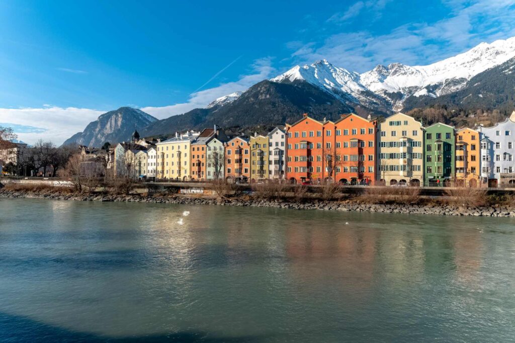 Eine Reihe farbenfroher Gebäude entlang eines Flusses mit Bergen im Hintergrund unter blauem Himmel, die den lebendigen Charme von Innsbruck einfangen - der Heimat der renommierten Kliniken für Osteopathie in Innsbruck.