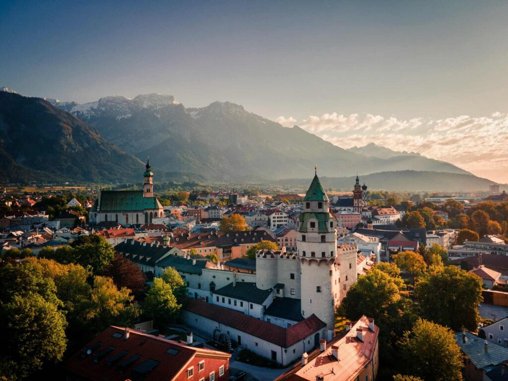 Luftaufnahme einer europäischen Stadt mit historischen Gebäuden, Kirchtürmen und umliegenden Bergen unter klarem Himmel, die den Charme von Innsbruck - der Heimat renommierter osteopathie innsbruck Kliniken - einfängt.